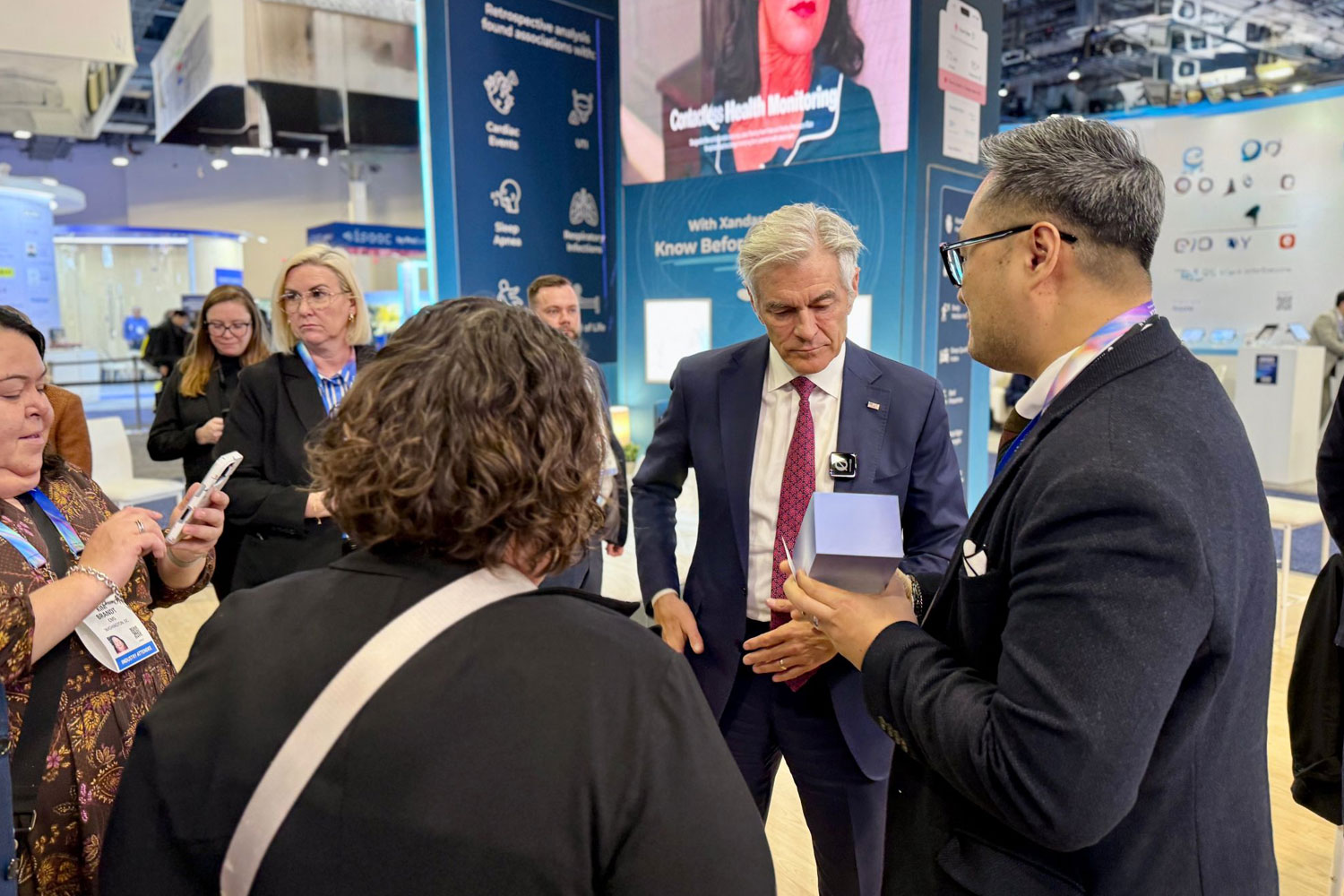 Federal healthcare leaders engage with Xandar Kardian on the CES show floor. A suited official examines a small medical technology device as company representatives explain its use. Attendees wearing conference badges look on, while large displays behind them highlight remote patient monitoring and vital-sign detection technologies focused on real-world healthcare system change.