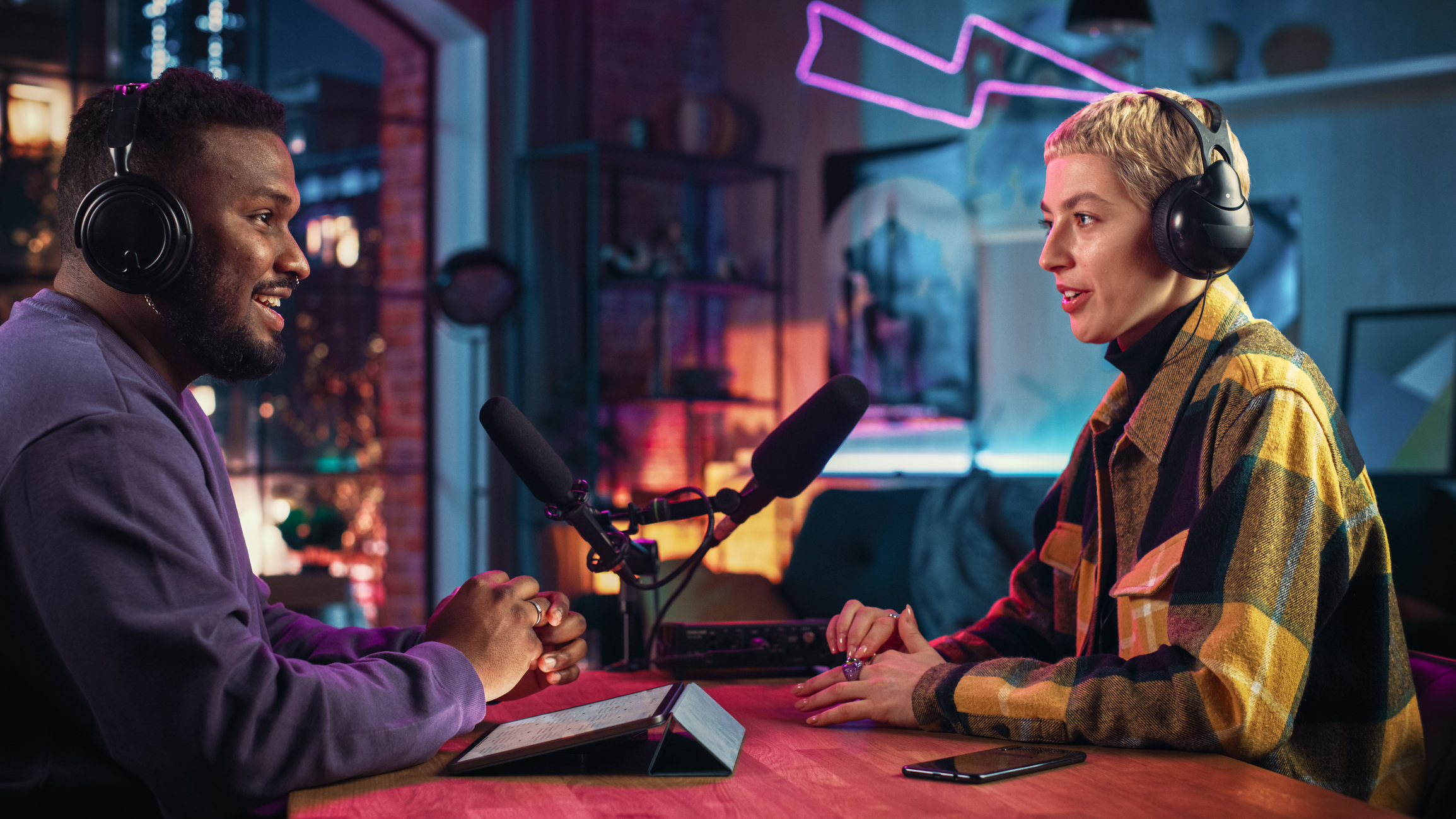 ALT Text: Two Young Stylish Radio Show Hosts Record Fresh Podcast Episode in Home Loft Studio Apartment. Attractive Energetic Co-hosts Discuss Important Topics Live on Air in an Evening Show.