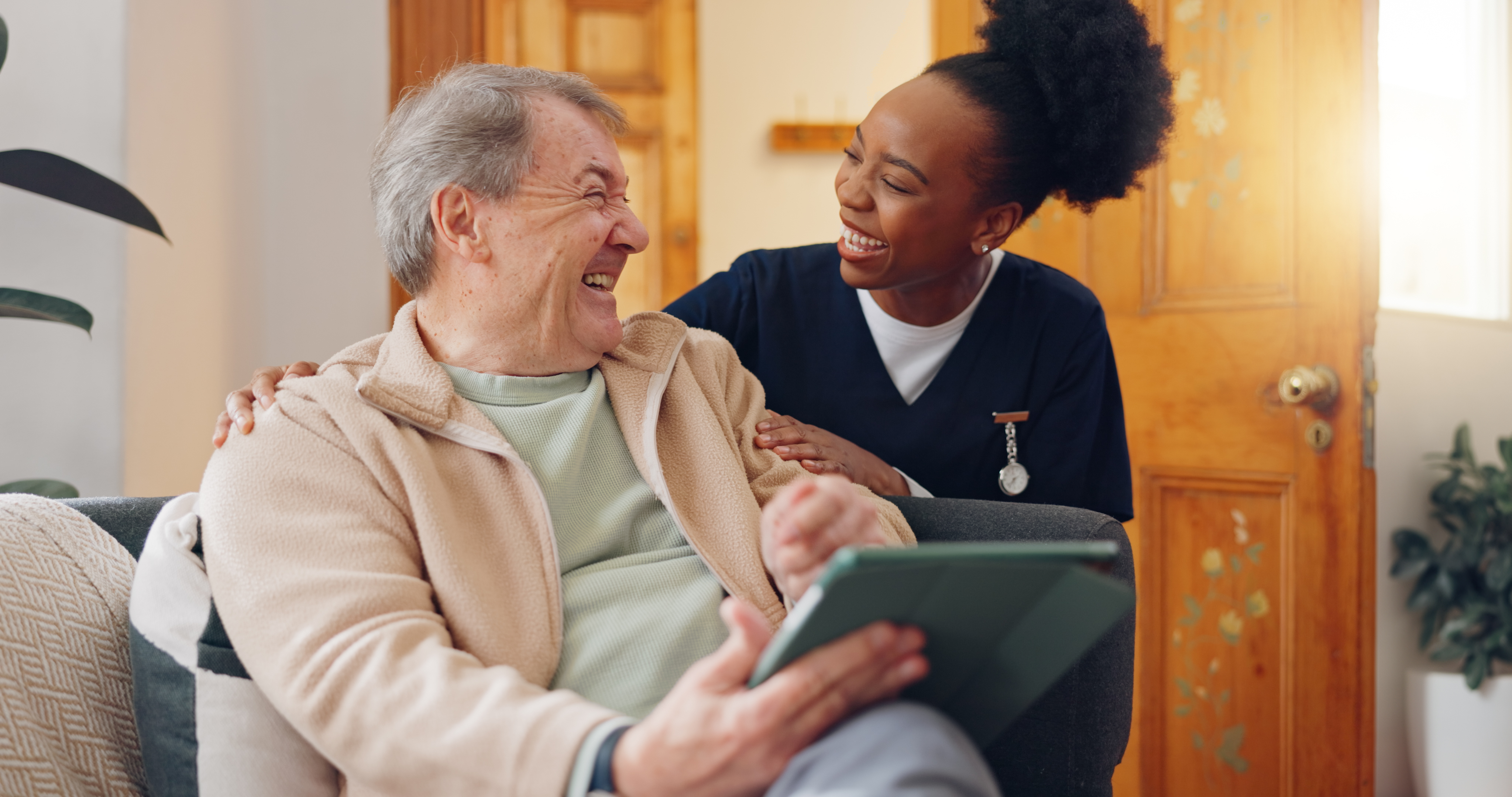 ALT Text: Smiling caregiver in scrubs interacting warmly with an elderly man holding a tablet.